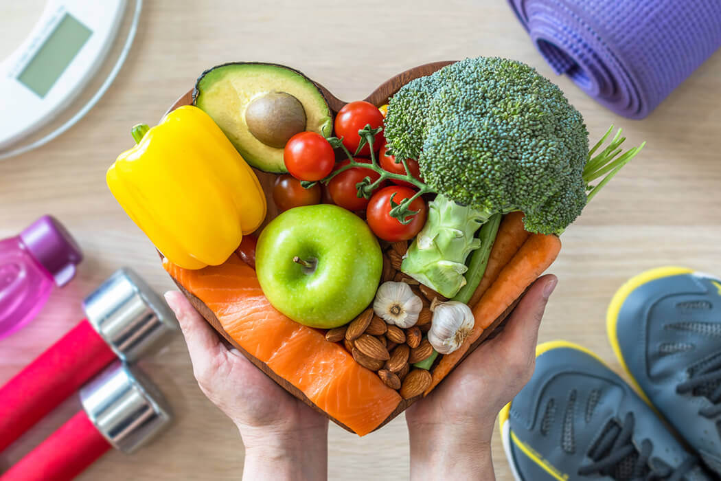 A heart-shaped wooden plate filled with heart-healthy foods including salmon, broccoli, a bell pepper, and small tomatoes.